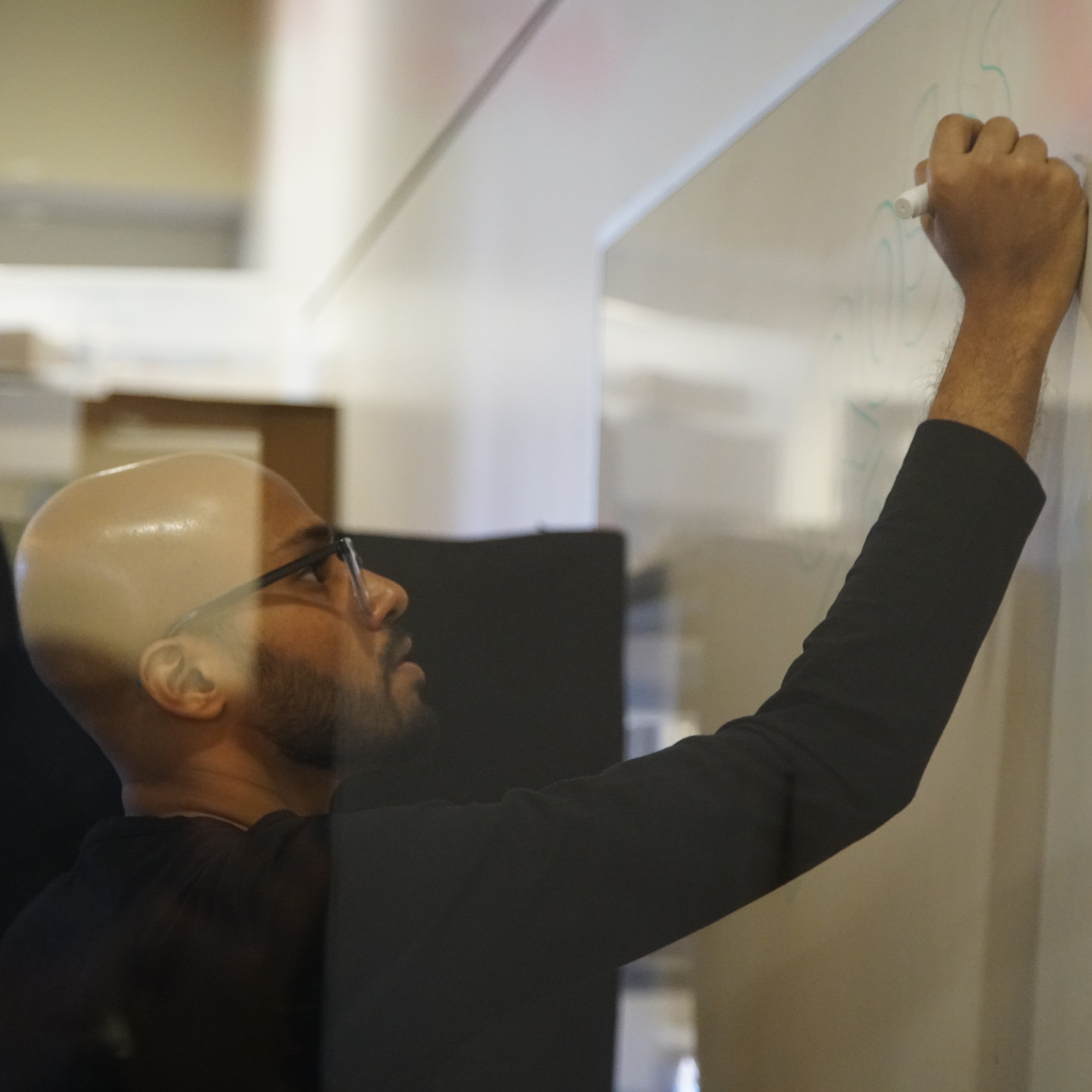 A photograph of Rishi drawing at a whiteboard. Rishi is a bespectacled man in his late-twenties, bald with brown skin and a trimmed beard and mustache. He is wearing a black full-sleeved t-shirt, and drawing on a whiteboard in profile.